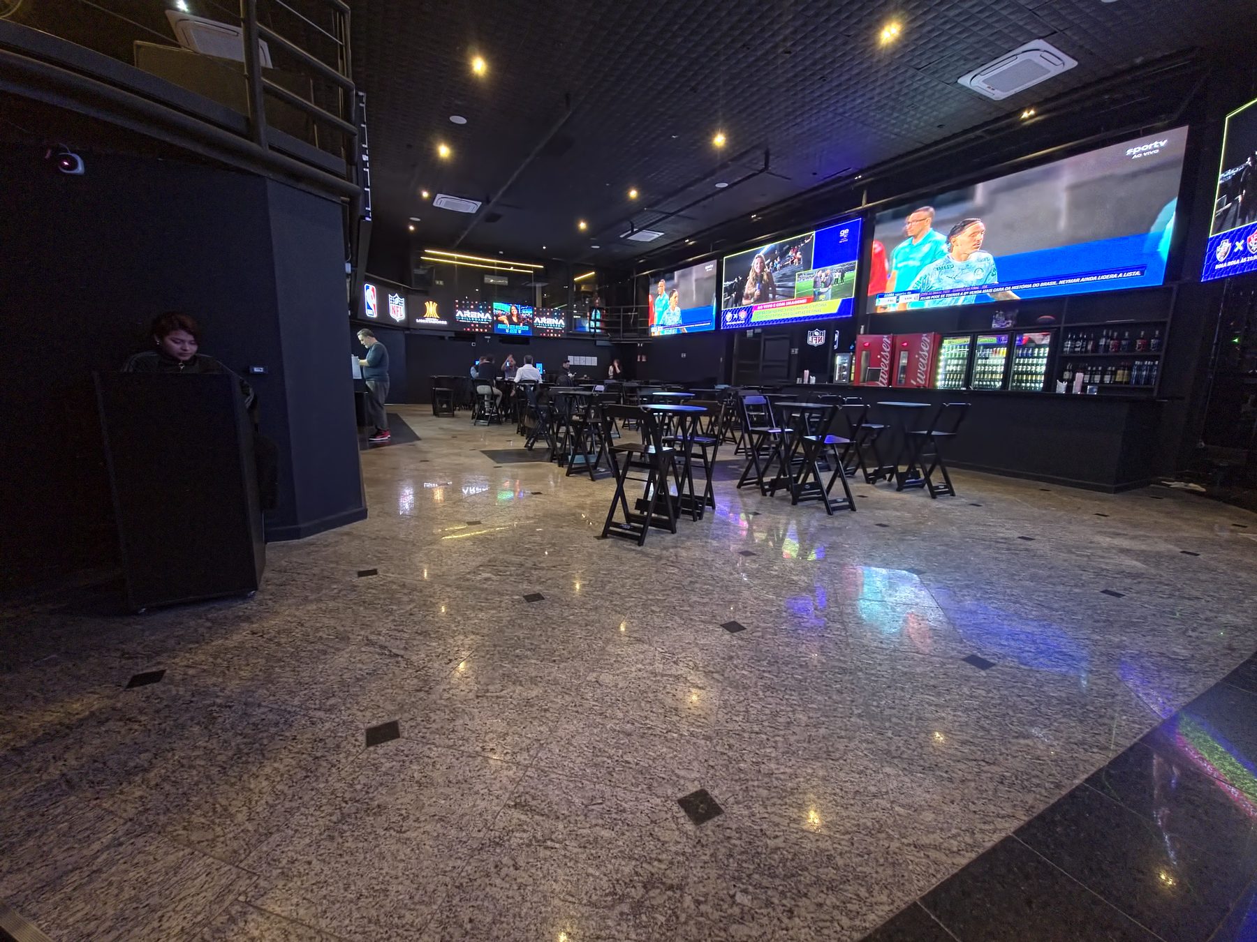 Área livre do térreo da Arena Billboard, com piso de granito, mesas altas junto ao bar e painéis de LED acesos ao fundo.
