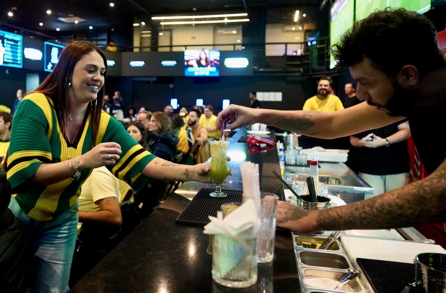 Bartender da Arena Billboard entregando um drink a uma cliente no balcão do bar, com o salão cheio ao fundo.