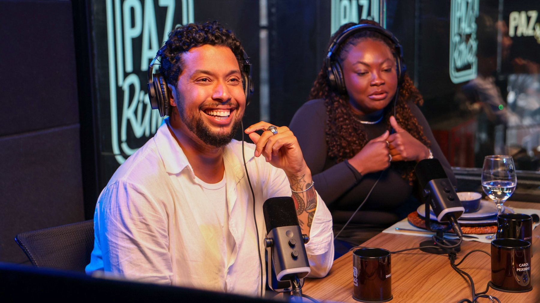 Gravação em andamento no estúdio de podcast da Arena Billboard: dois apresentadores com fones de ouvido diante de microfones, na bancada de madeira do set.