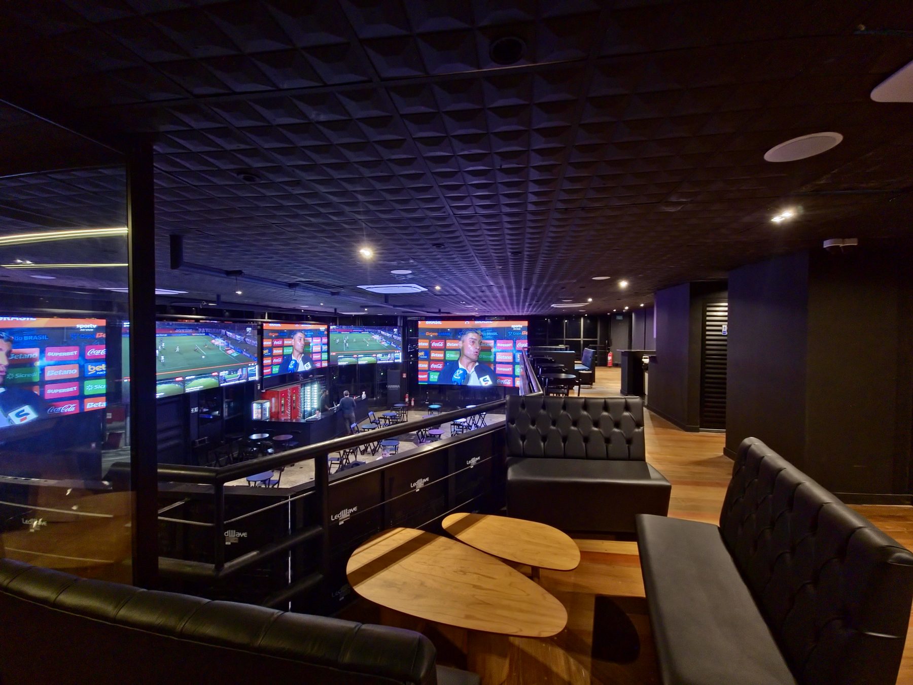 Lounge do mezanino da Arena Billboard, com sofás de couro, mesas baixas de madeira e vista para o salão e os painéis de LED.