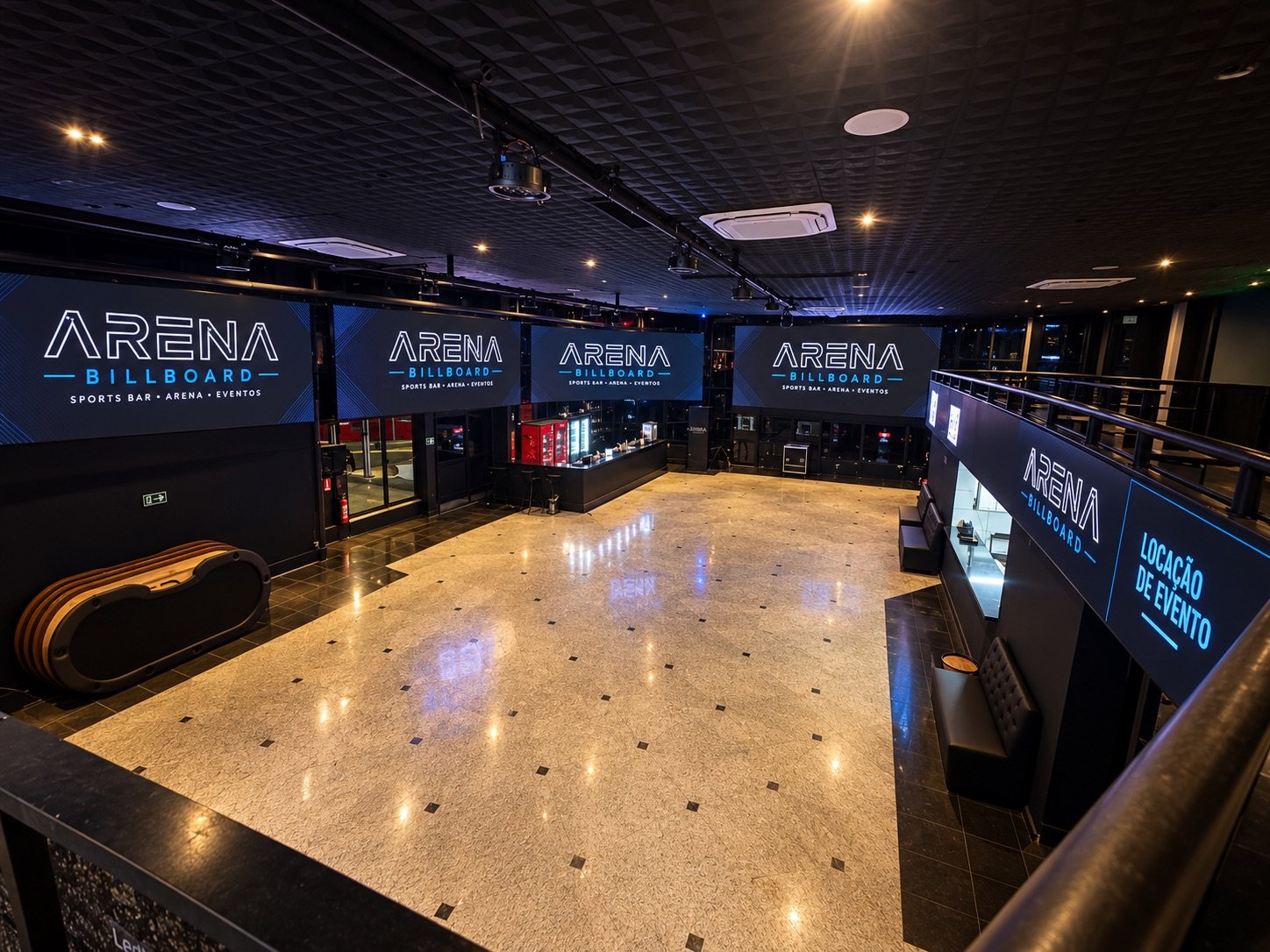 Salão principal da Arena Billboard vazio, visto do mezanino: piso de granito polido, bar ao fundo e quatro painéis de LED exibindo a marca da casa.